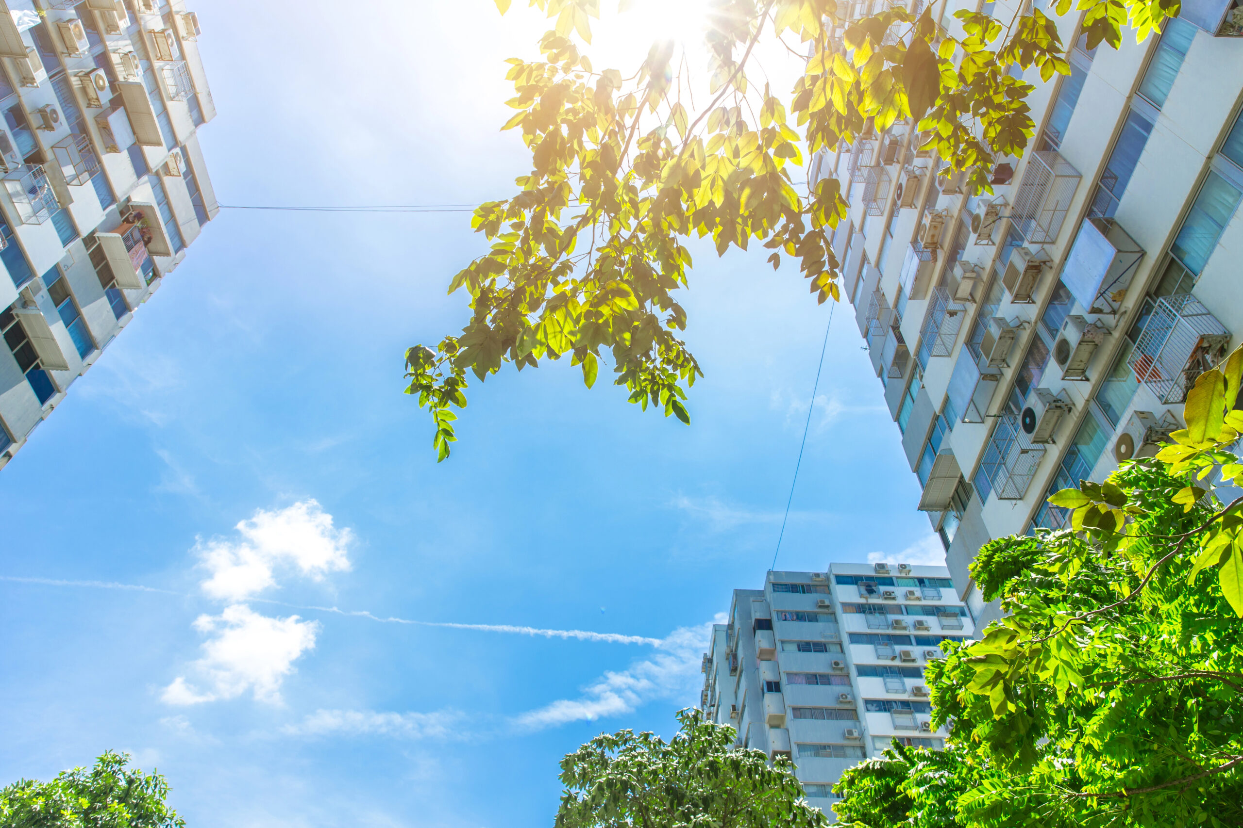Zonnige dag tussen twee flatgebouwen met groene bomen en zicht op aircosystemen aan de gevels.